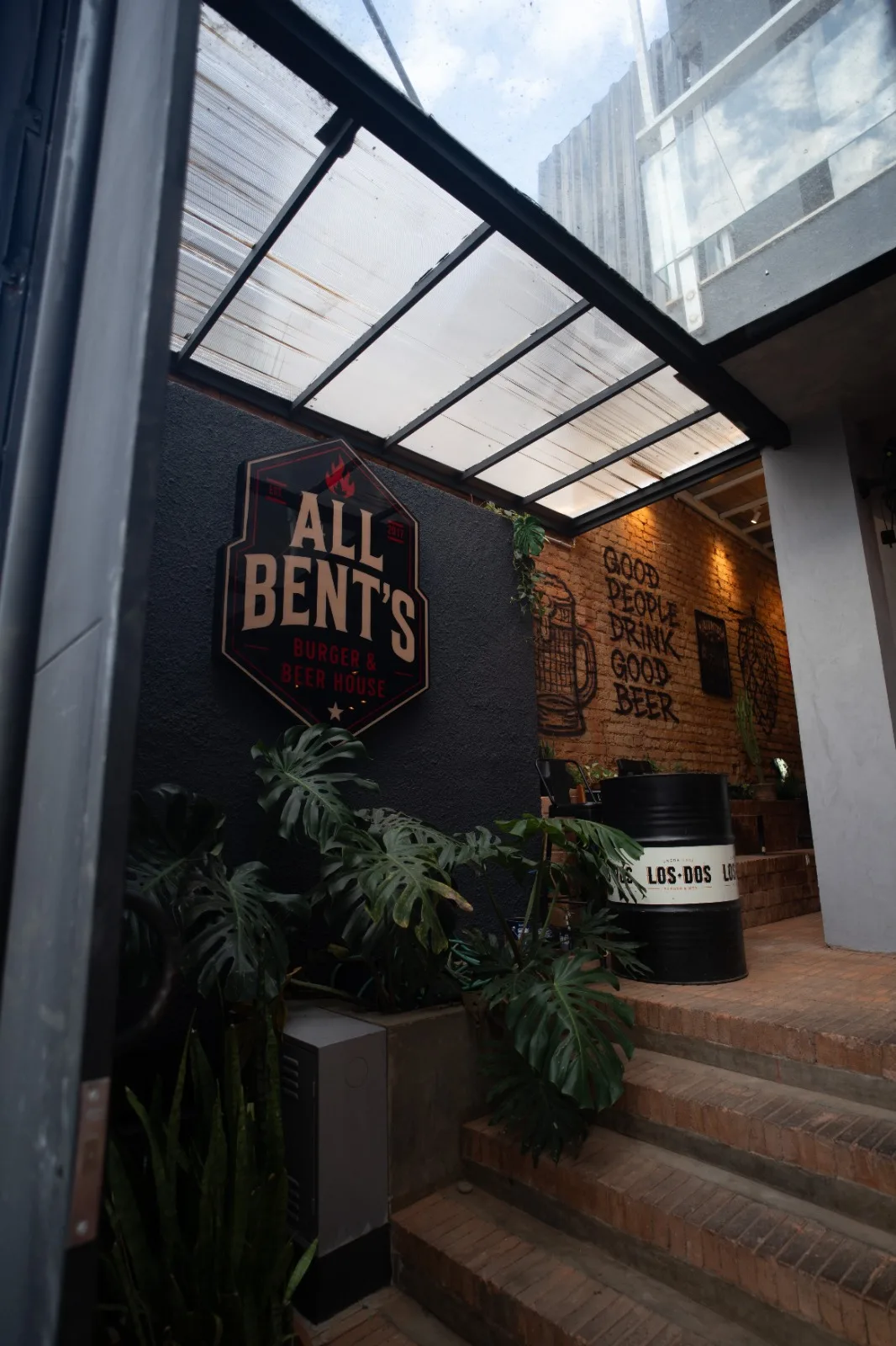 Entrada interna da All Bent's com parede de tijolos, plantas e identidade da casa